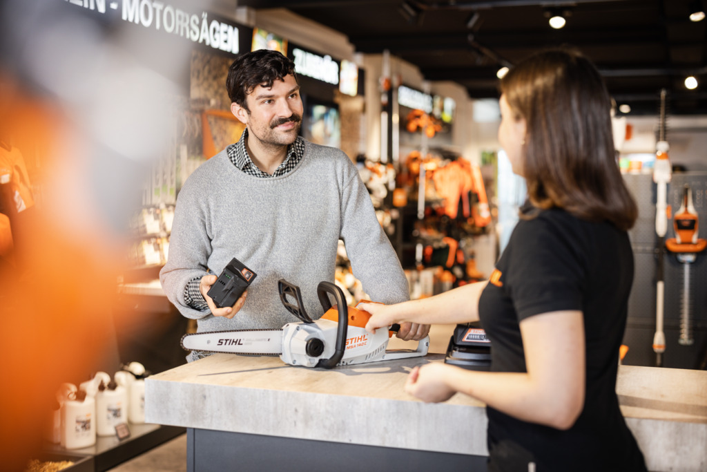 A man in a gray sweater holds a battery while talking to a woman at a hardware store counter with a STIHL chainsaw between them.