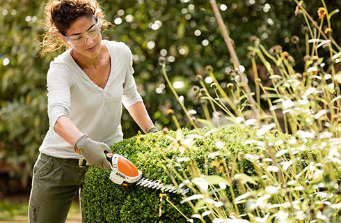A woman wearing safety glasses and gloves trims a hedge into shape using STIHL HSA 25 cordless shrub shears