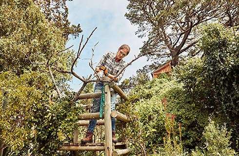 A woman wearing safety glasses and gloves stands on a wooden scaffold cutting a branch with the STIHL GTA 26 cordless garden pruner