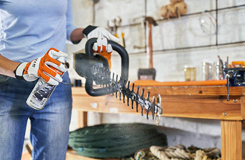 A STIHL HSA 56 cordless hedge trimmer is held up in one hand by a woman wearing gloves, who is spraying resin solvent onto the cutting apparatus with the other hand