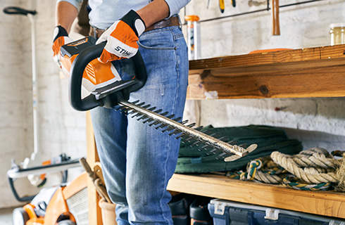 A STIHL HSA 56 cordless hedge trimmer being held by someone wearing gloves, in front of a workbench and lawn mower