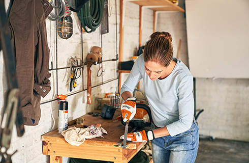 A woman wearing protective gloves using a flat file and sharpening a hedge trimmer on a wooden workbench