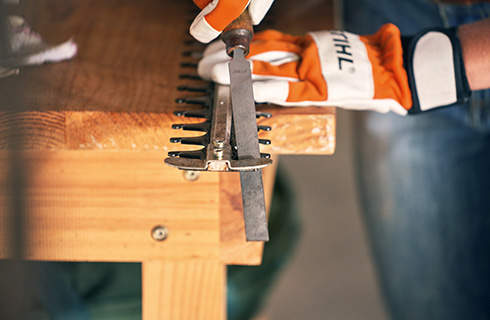 Close-up of a flat file being used to sharpen one blade of a hedge trimmer that overhangs a workbench, with gloved hands visible in the background