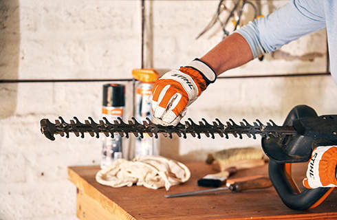 A person wearing gloves holds a hedge trimmer on a workbench while using a cloth to clean the cutting apparatus
