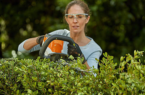 A woman wearing eye protection and protective gloves, using a hedge trimmer to cut the top of a green hedge that she is standing behind