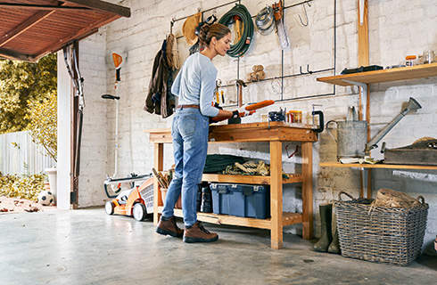 A woman wearing protective gloves stands at a workbench in a tidy garage, working on a STIHL HSA 56 cordless hedge trimmer