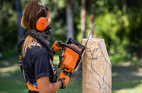 A woman wearing ear protection, gloves and a bandana over her mouth using a STIHL chainsaw to roughly carve a piece of wood with an owl drawn on it.