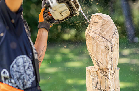 A roughly carved wooden owl has details added to its face by a woman using a STIHL chainsaw with a carving set.