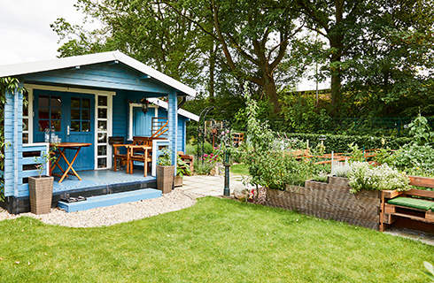 Garden with green lawn in the foreground, bordered by a blue garden building and a stepped raised bed with a bench.