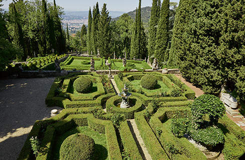 View over a formal topiary garden containing geometric hedging borders and topiary balls, with tall cypress trees in the background.