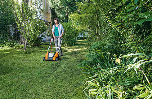 A woman uses a STIHL RLE 240 electric lawn aerator to scarify a lawn that contains several shrubs and trees