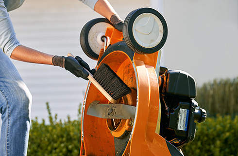 View of the underside of a lawn mower that is being held upright by a person wearing gloves and using a brush to clean near the blade