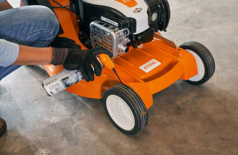 A lawn mower has lubricant spray applied to a front wheel by a person wearing gloves