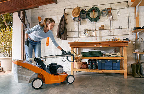 A woman wearing gloves folds down the handlebar of a lawn mower inside a tidy garage, with a workbench behind