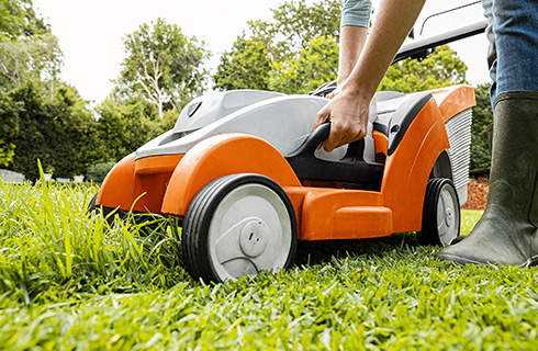 A STIHL RMA 339 cordless lawn mower on a partly mown lawn, as someone leans over it to use the control panel