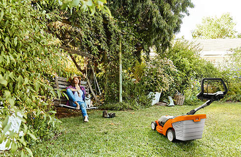 A woman relaxes on a garden swing next to a STIHL RMA 339 cordless lawnmower on a green plot