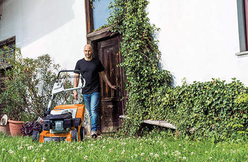 An unmown lawn outside a white building, from which a man walks towards a STIHL RM 650 V petrol mulching mower