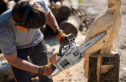 A man wearing ear protection uses a STIHL MS 193 C-E with carving attachment to cut feathers into an eagle sculpture
