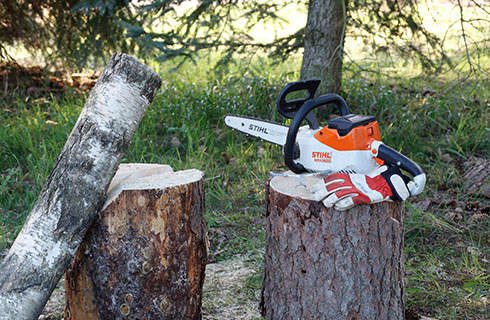 A STIHL MSA 140 C chainsaw and protective gloves on a tree stump with other logs nearby