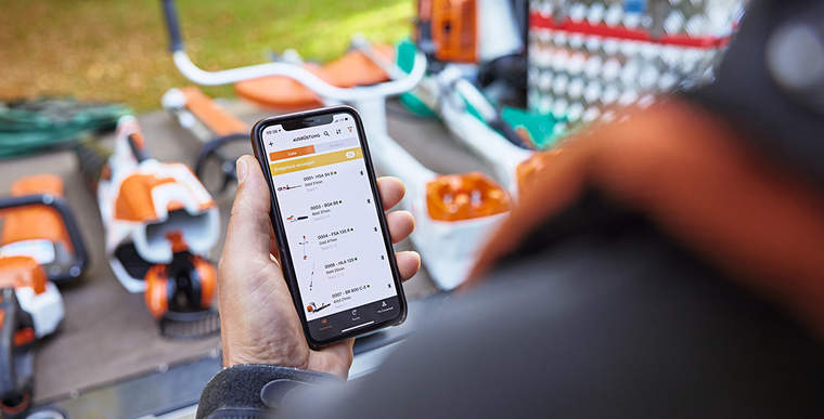 A person standing in front of the loading area of a platform vehicle on which various STIHL tools are laid out. The person holds a smartphone in their hand with the STIHL connected App open.