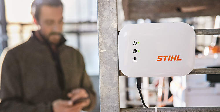 A STIHL connected Box hung on a workshop shelf. In the background there is a gardener and landscaper interacting with their smartphone.